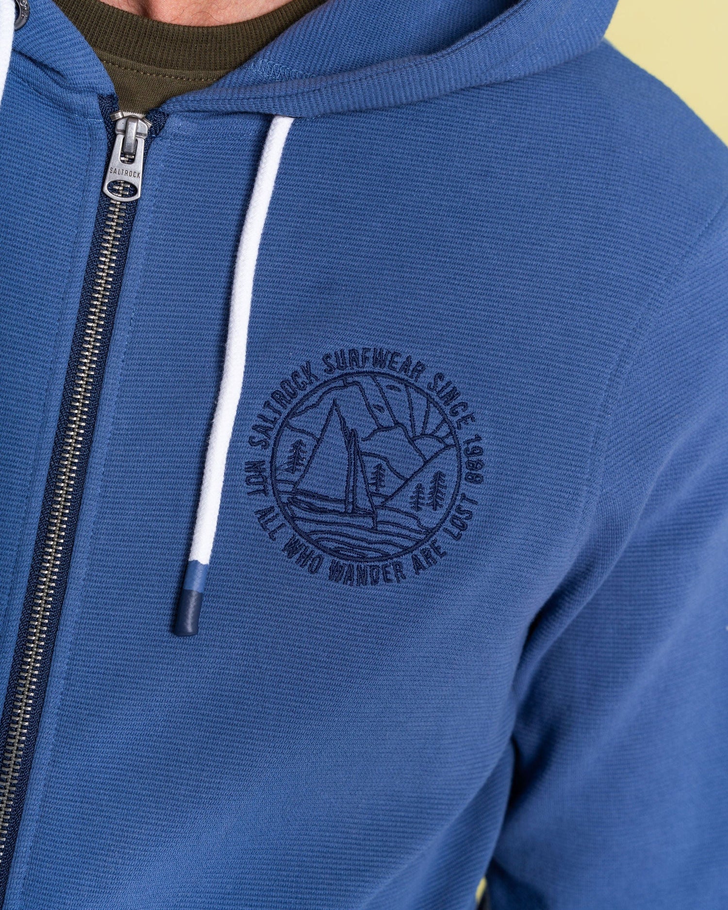 A person is wearing the Hanns - Mens Zip Hoodie by Saltrock, featuring white drawstrings and their iconic logo of mountains and a boat. Made from 100% cotton, this blue hoodie captures the spirit of "Not All Who Wander Are Lost.