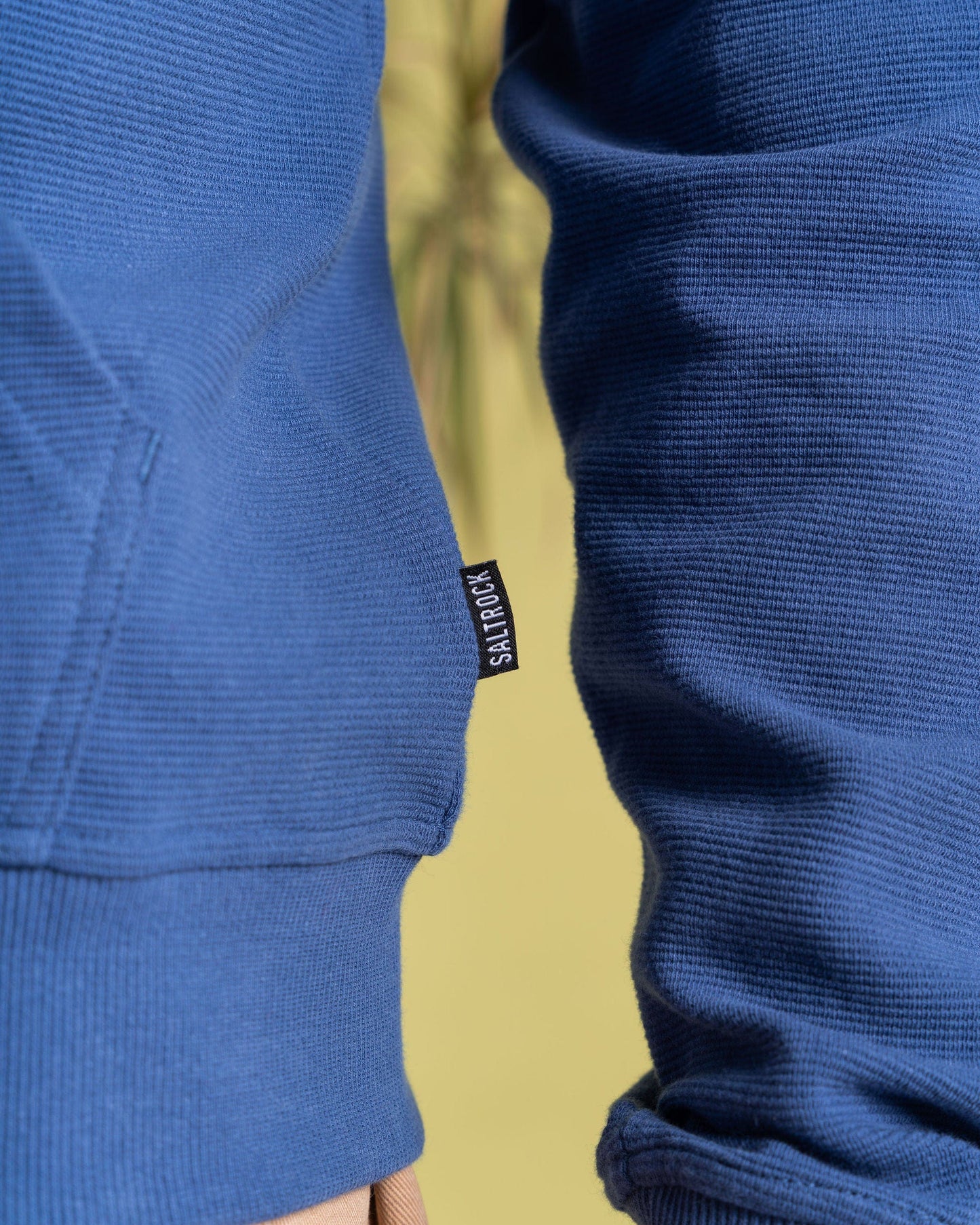 Close-up of someone wearing a Saltrock Hanns Men's Zip Hoodie in blue with a small "SHADYBOX" tag. Made from 100% cotton, it has a subtle ribbed texture. Background is light yellow-green.