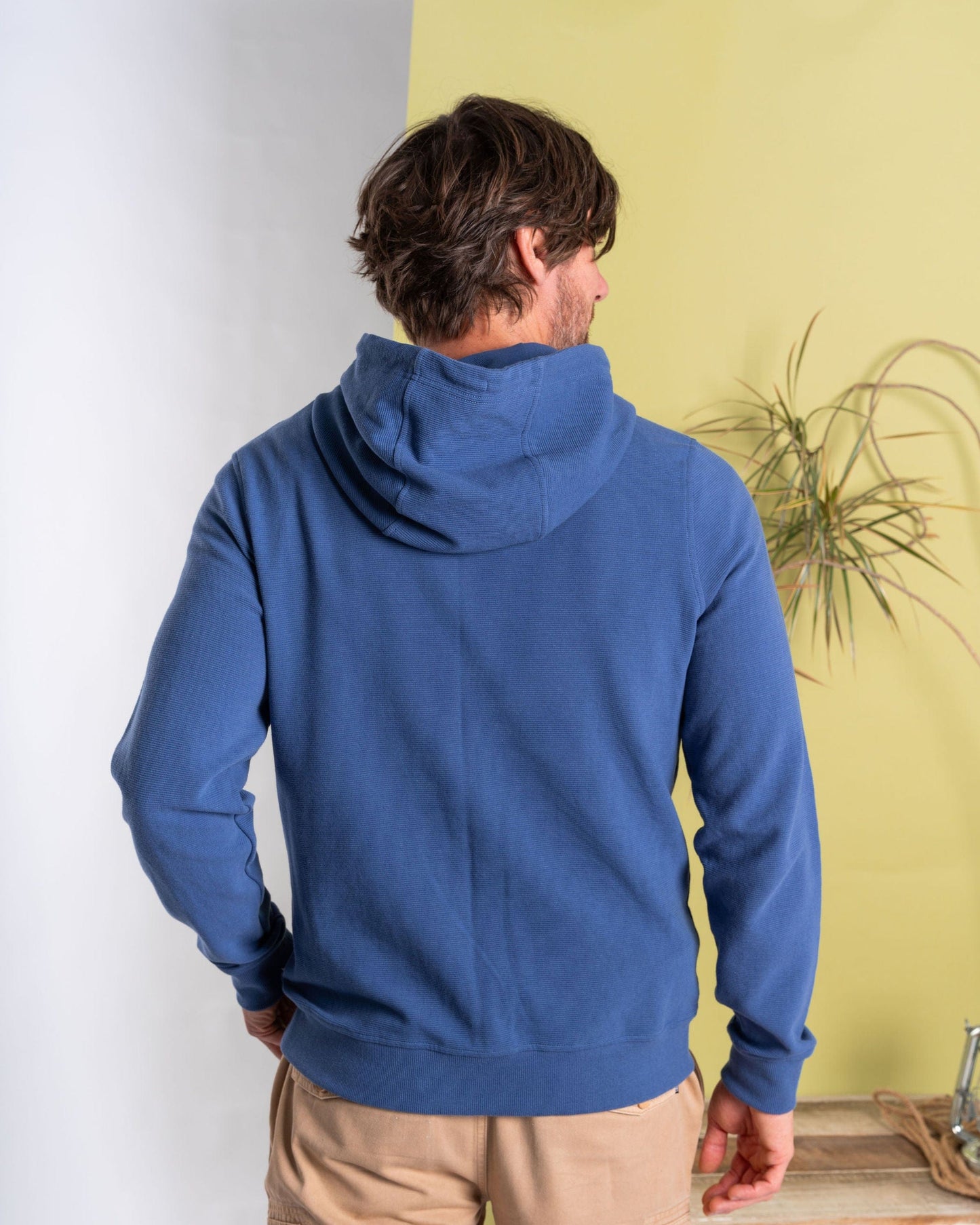 A person with medium-length hair faces away in a Saltrock branded blue "Hanns - Mens Zip Hoodie," paired with beige pants. They stand against a light green wall, near a plant and wooden furniture, creating a cozy vibe.