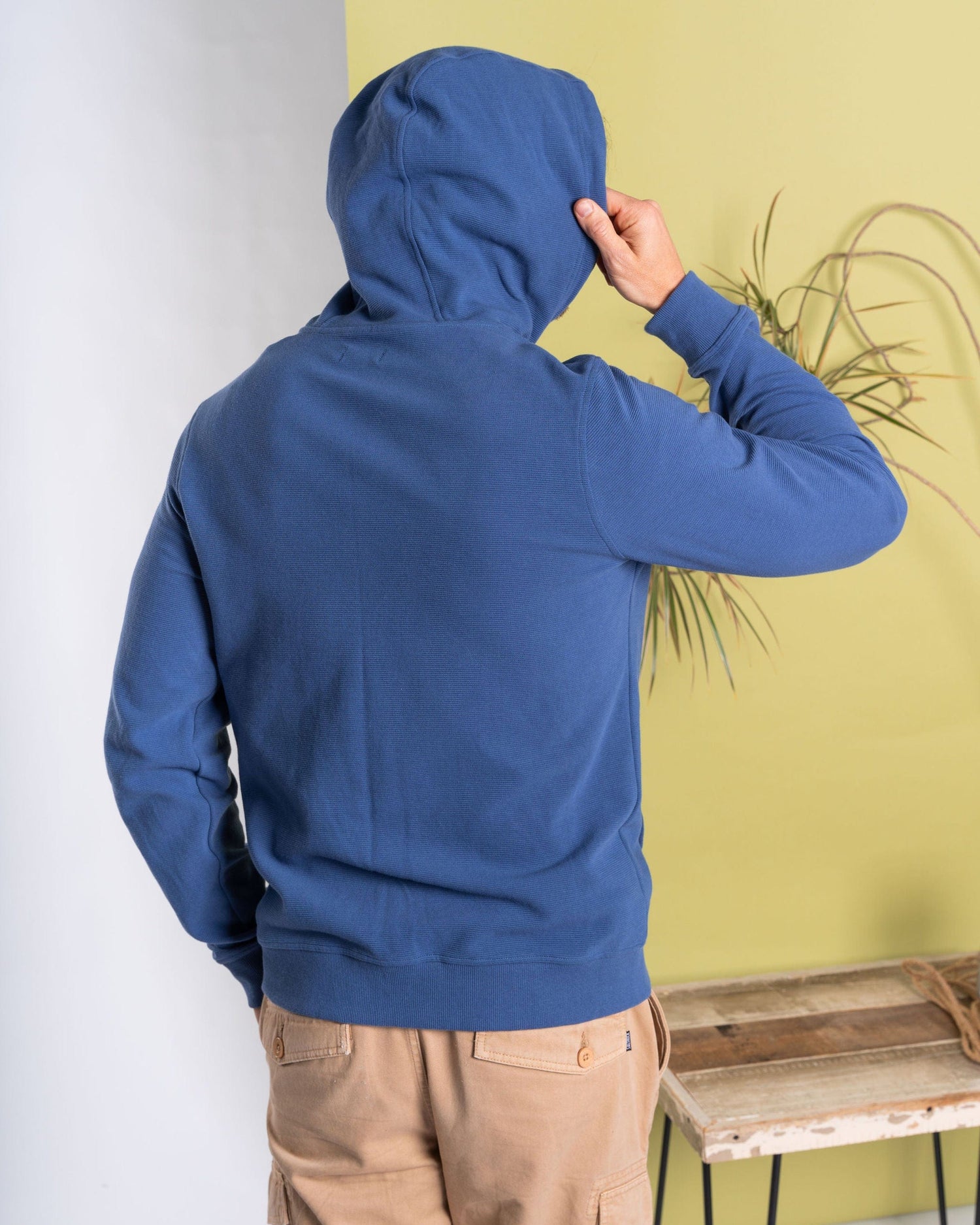 Wearing the Hanns - Mens Zip Hoodie in blue by Saltrock, a person stands with the hood up against a green and white wall, while a nearby plant adds natural flair to the scene.