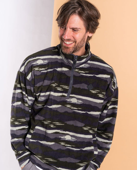 A man in the Saltrock Hawker oversized green fleece pullover made from recycled materials stands against a two-tone background, gazing down and smiling.