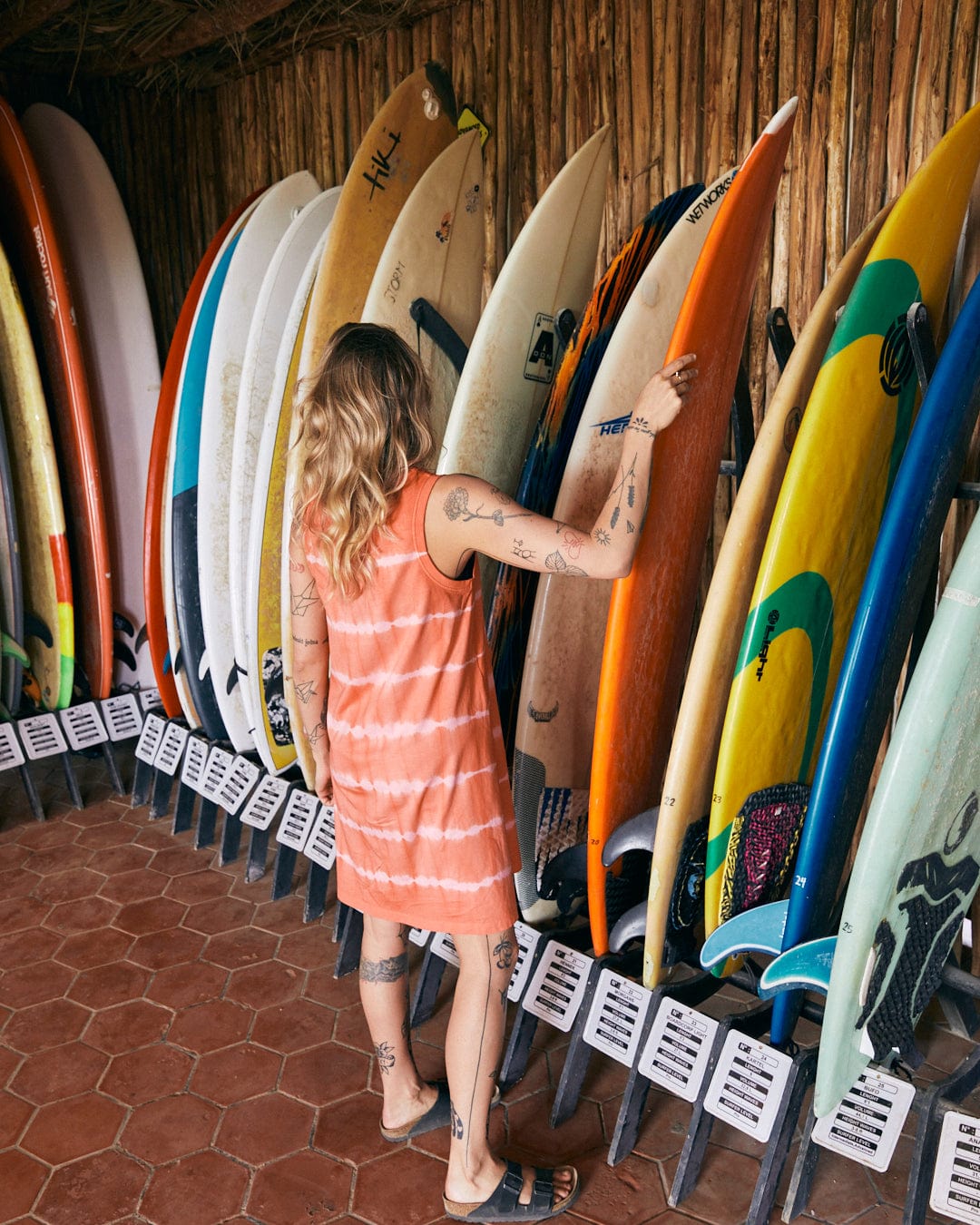 A person with tattoos and long hair in an Eliana - Womens Tie Dye Vest Dress - Peach by Saltrock stands in front of a rack of surfboards, touching one of them in a rustic, wood-paneled room.