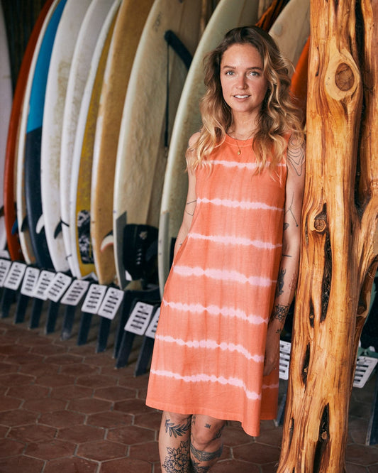 A woman in an Eliana - Womens Tie Dye Vest Dress - Peach with embroidered Saltrock branding stands by a wooden post, with a row of surfboards behind her.