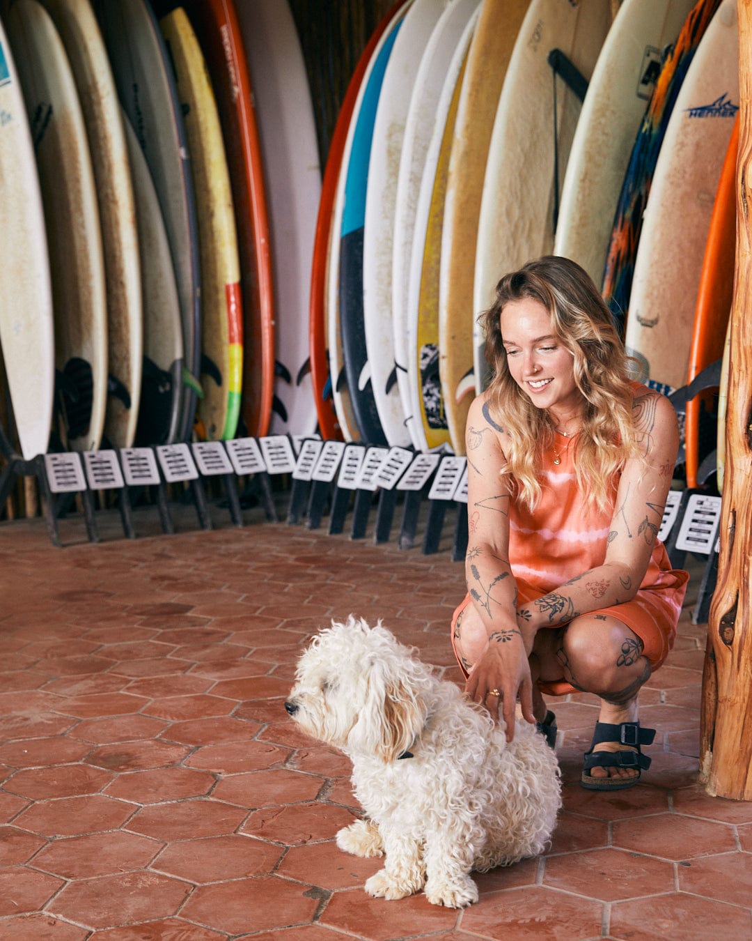 A person in a Saltrock Eliana - Womens Tie Dye Vest Dress - Peach and sandals is petting a fluffy white dog in front of a display of surfboards.