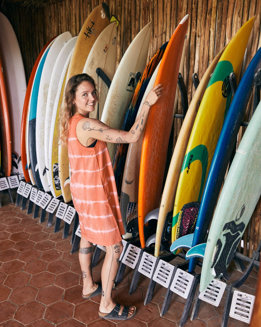 A person wearing an Eliana - Womens Tie Dye Vest Dress - Peach from Saltrock and sandals smiles while standing next to a rack of surfboards, reaching for an orange one.