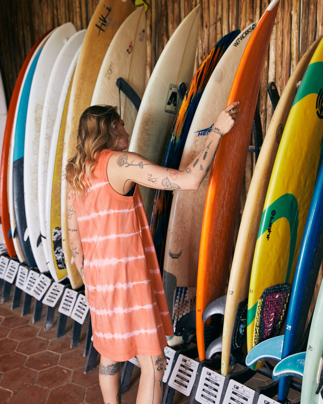 A person in a Saltrock Eliana - Womens Tie Dye Vest Dress - Peach is browsing through a rack of surfboards mounted against a wall.