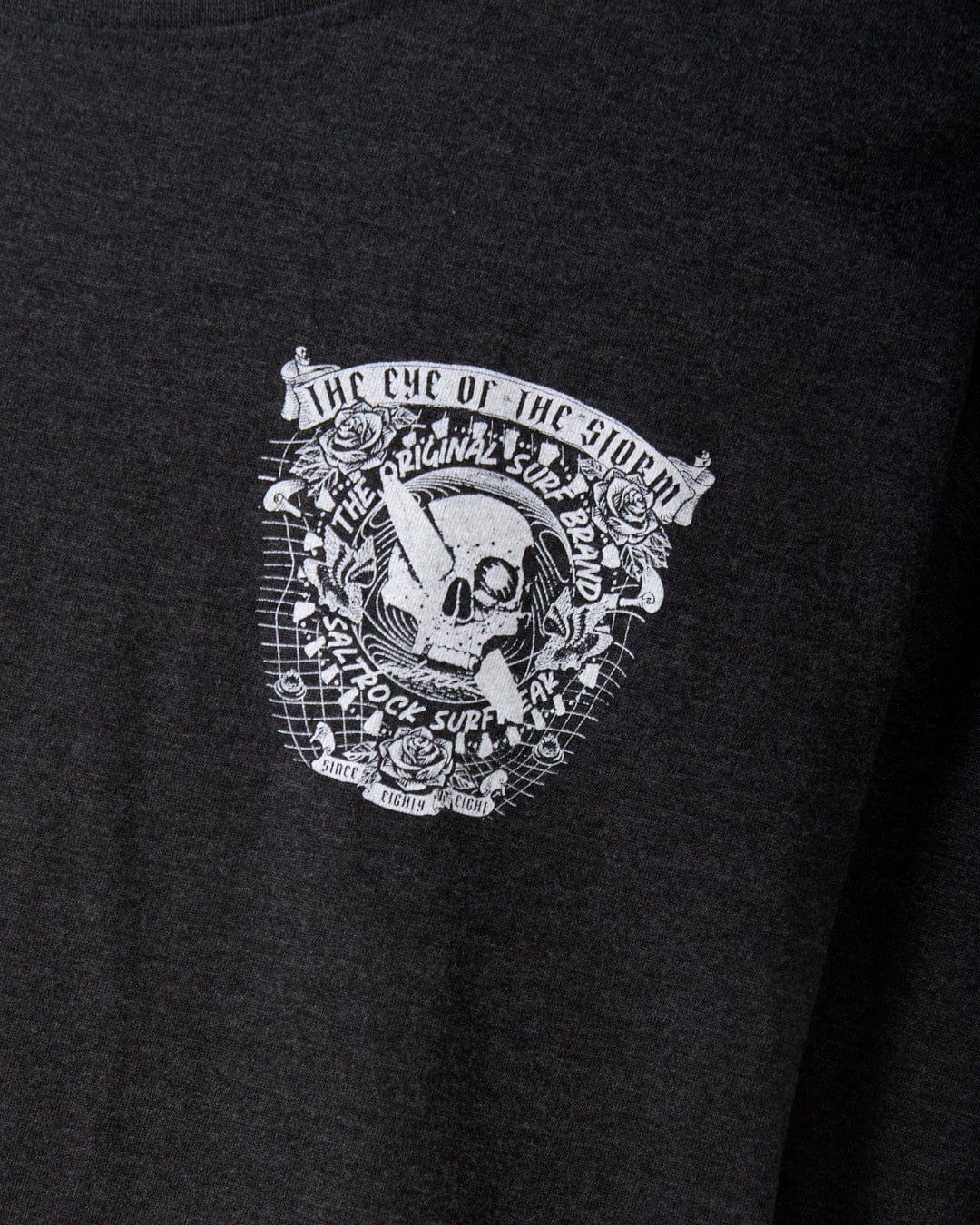 Detailed view of a grey long sleeve T-shirt from Saltrock, featuring a striking white skull and surf graphic design. It prominently showcases the phrases "The Eye of the Storm," "The Original Surf Band," and "Hillrock Supply Co." A classic crew neckline makes this an essential piece for your wardrobe.