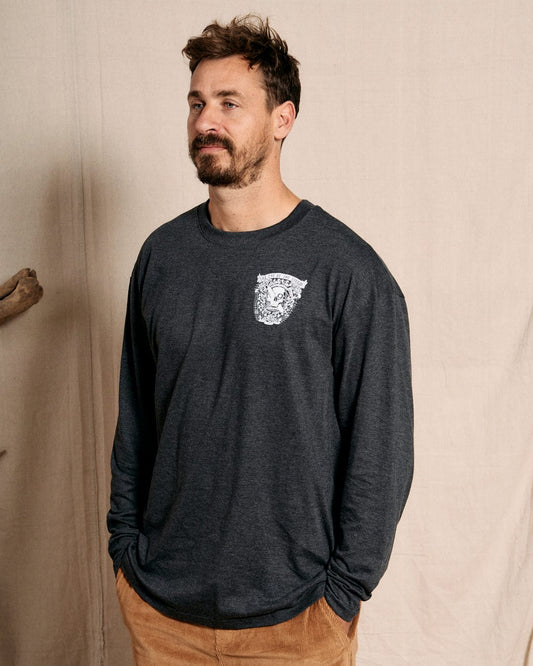 A man with a short beard is wearing the Eye Of The Storm 2 Saltrock long-sleeve t-shirt in grey, featuring a small skull graphic on the chest and a classic crew neckline, standing against a plain beige background.