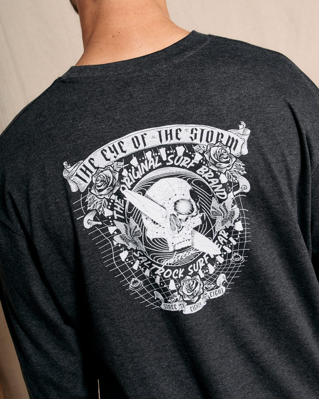 A back view of someone wearing the "Eye Of The Storm 2" men's long sleeve t-shirt in grey showcases an edgy design with skull and surf graphics, roses, and the text "The Eye of the Storm." This Saltrock shirt, promoting the brand's surf and rock fair theme, features a crew neckline that perfectly complements its bold look.