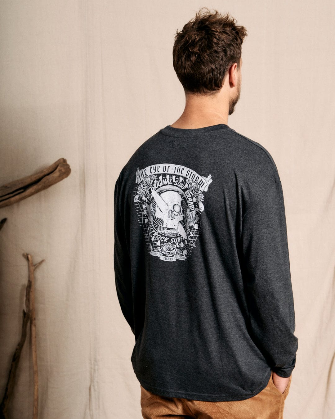 A person with short hair faces away from the camera, wearing a grey long-sleeve shirt from Saltrock's "Eye Of The Storm 2" collection, featuring a skull/surf graphic design and a crew neckline.