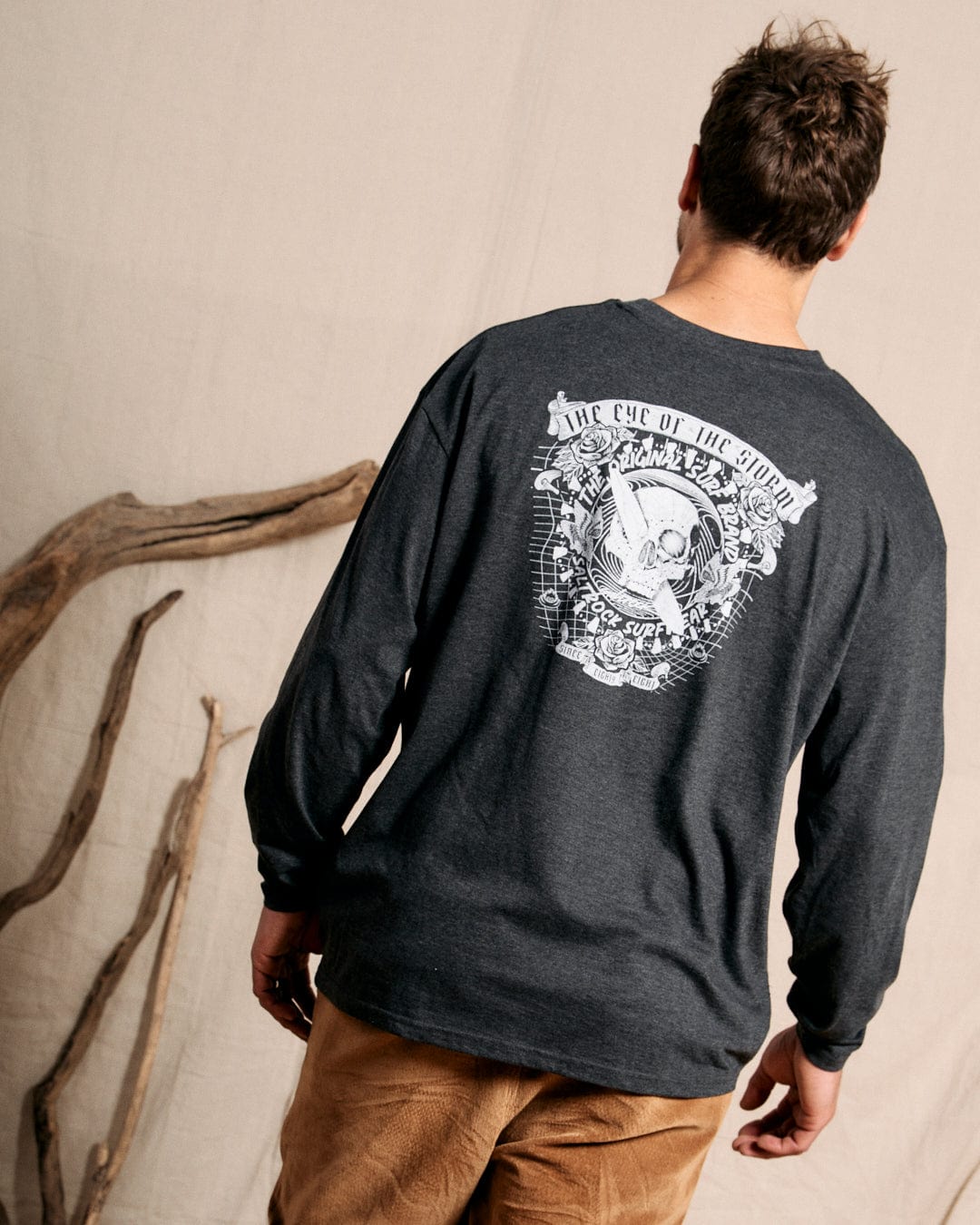 A person wearing the Eye Of The Storm 2 men's long-sleeve T-shirt in grey, featuring intricate skull graphics on the back, stands near a few wooden branches against a neutral backdrop. The Saltrock branding complements the crew neckline, creating a balanced and stylish look.