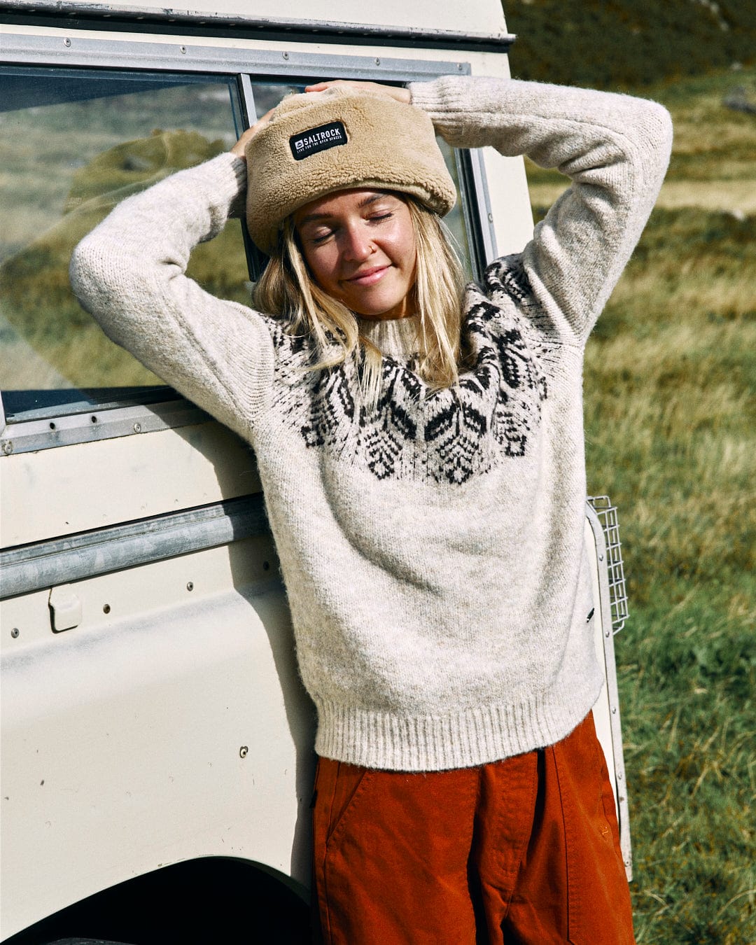 A person in the Saltrock Fairisle Women's Knitted Jumper and a beanie leans against a vehicle with eyes closed in a grassy area.