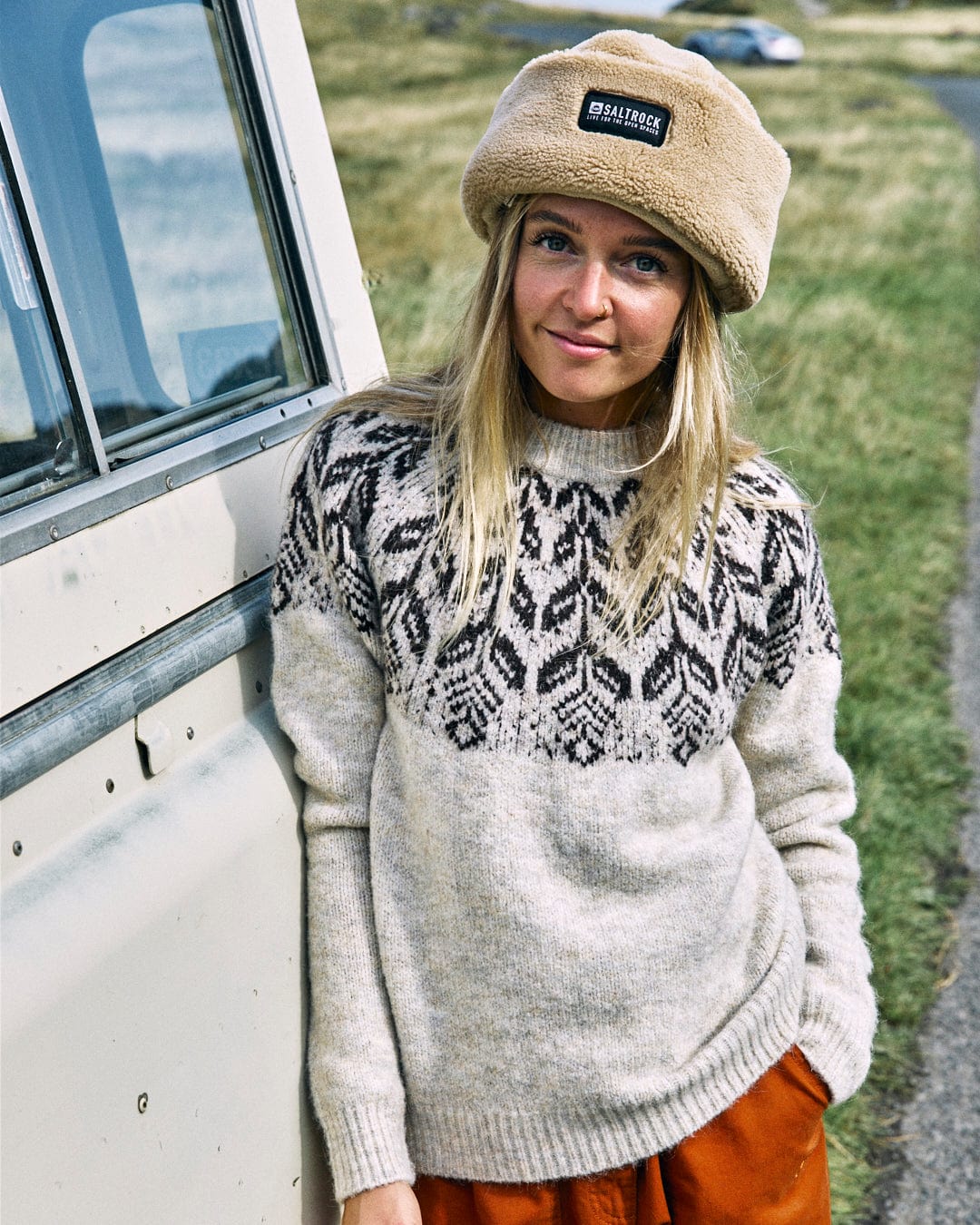 A person wearing the Saltrock Fairisle Women's Knitted Jumper and a fur hat leans against a vehicle, standing outdoors with grass in the background.