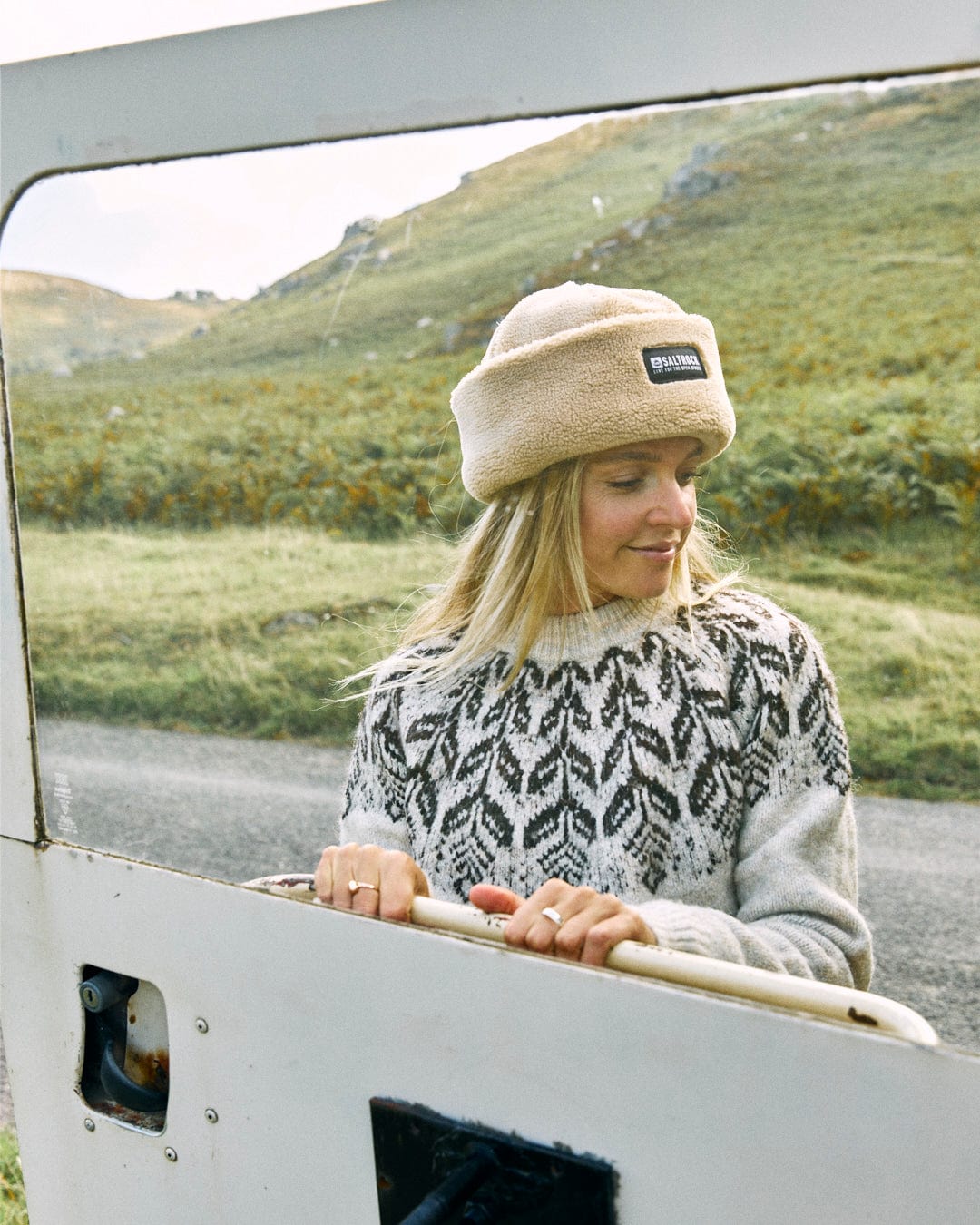 A woman in a machine washable patterned Saltrock Fairisle Women's Knitted Jumper and a beige hat leans on a car door, facing down, with a grassy hillside and road in the background.