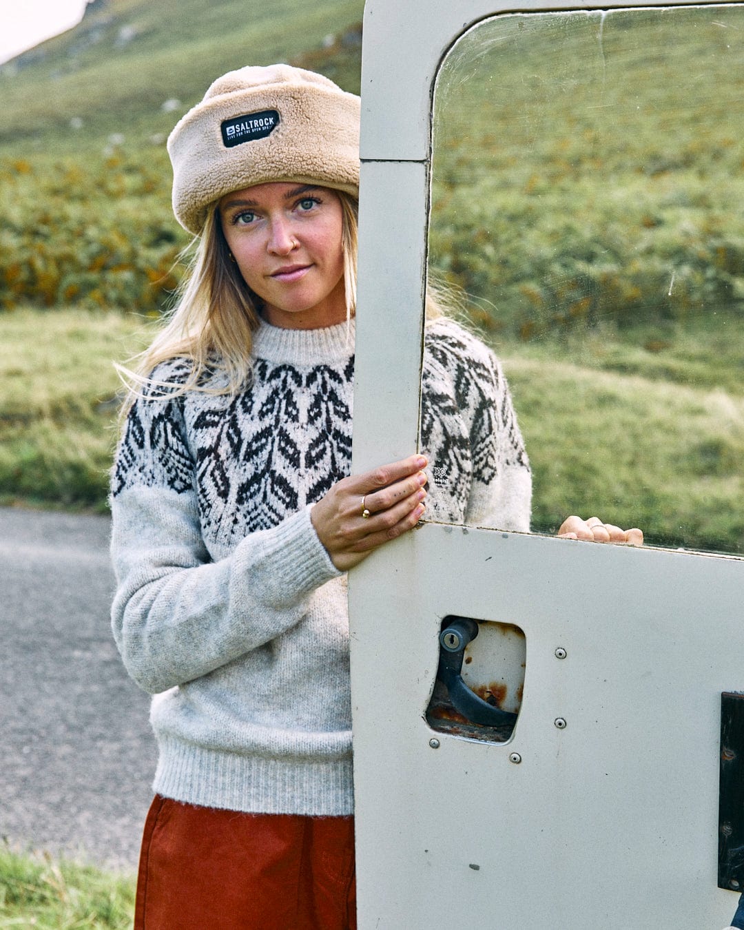 A woman wearing the Fairisle - Women's Knitted Jumper by Saltrock and a fur hat stands by an open car door in a grassy landscape.