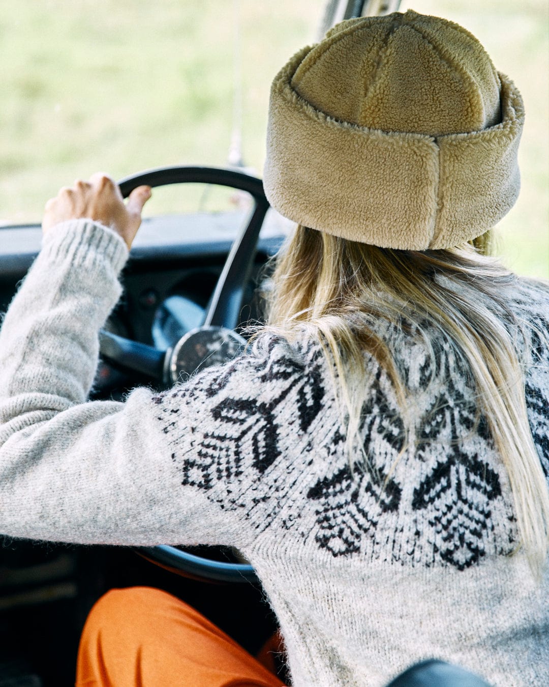 Person wearing a Fairisle women's knitted jumper from Saltrock and a beige fur hat drives a vehicle, viewed from behind.