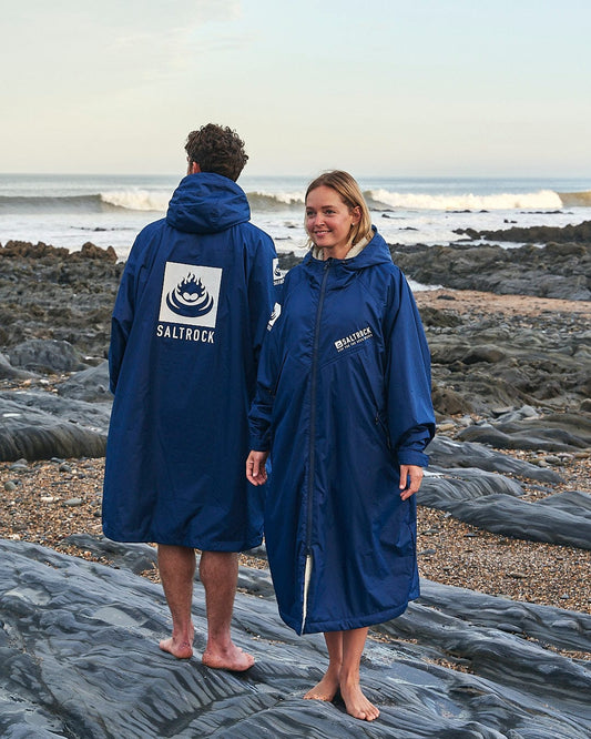 Two people stand on a rocky beach, wearing blue Saltrock branded hooded robes. The person facing forward smiles while the other looks towards the ocean. Waves are visible in the background. The unmistakable Saltrock logo adds a touch of authenticity to their Changing Robe - Blue, perfect for any weather.