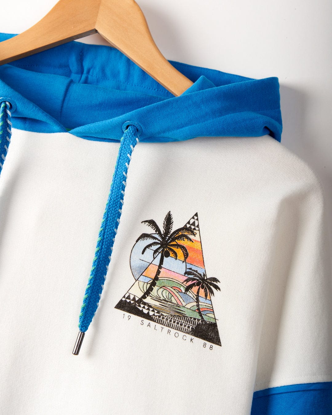 A white and blue hoodie on a hanger, featuring Palm Beach graphics of a tropical sunset scene with palm trees and waves. Text below the graphic reads "19 Saltrock 88." Crafted from 100% cotton, this Geo Beach - Womens Pop Hoodie - White also showcases signature Saltrock branding.