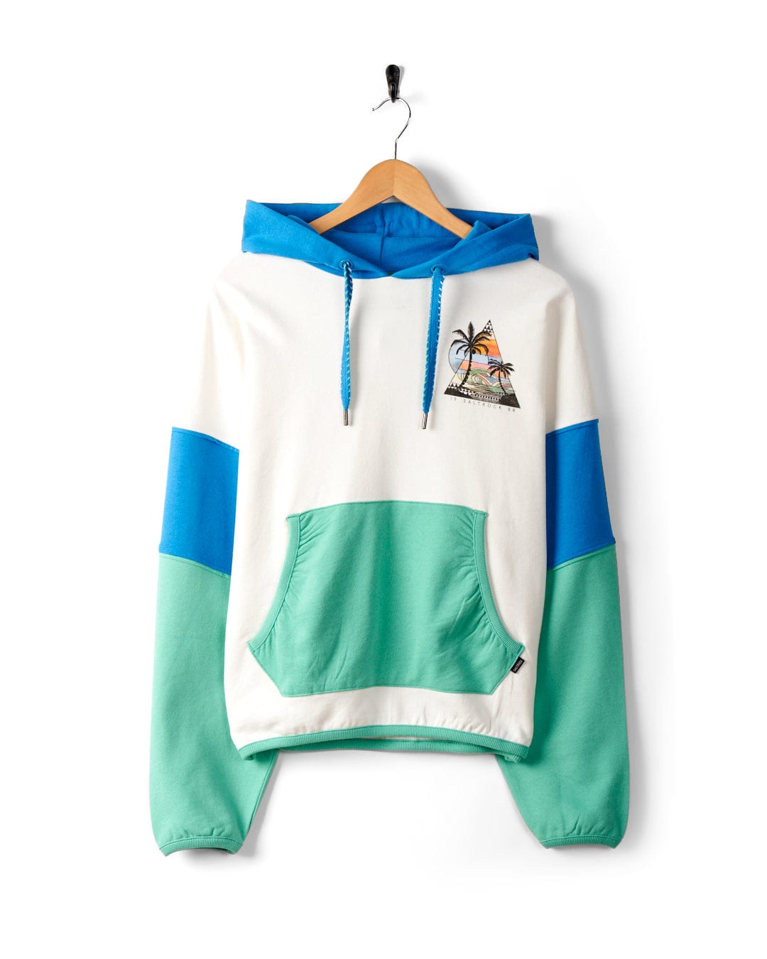 A hooded sweatshirt with white, blue, and green color blocks hangs on a wooden hanger. The Geo Beach - Womens Pop Hoodie - White, made of 100% cotton, features a front pocket and an embroidered palm tree and sunset design on the chest with subtle Saltrock branding.