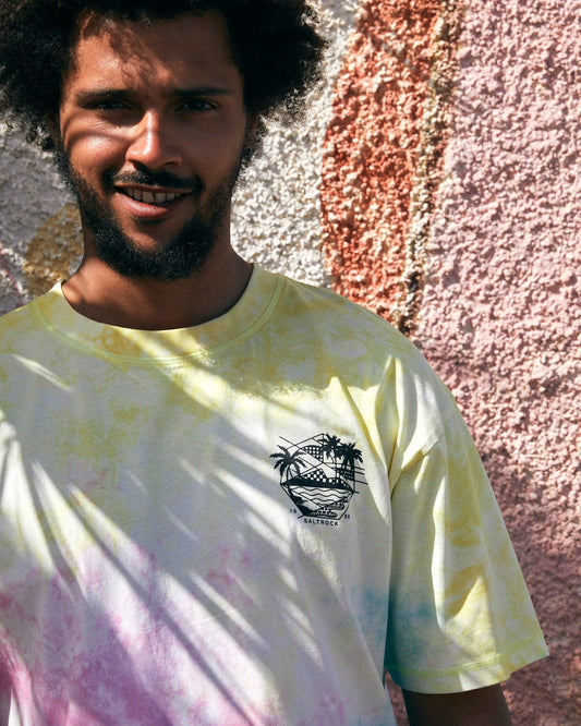 A man with curly hair and a beard stands in front of a textured wall, wearing the Saltrock Geo Palms Men's Tie Dye T-Shirt in Multi, an oversized fit made from 100% cotton, featuring vibrant palm trees and a graphic.
