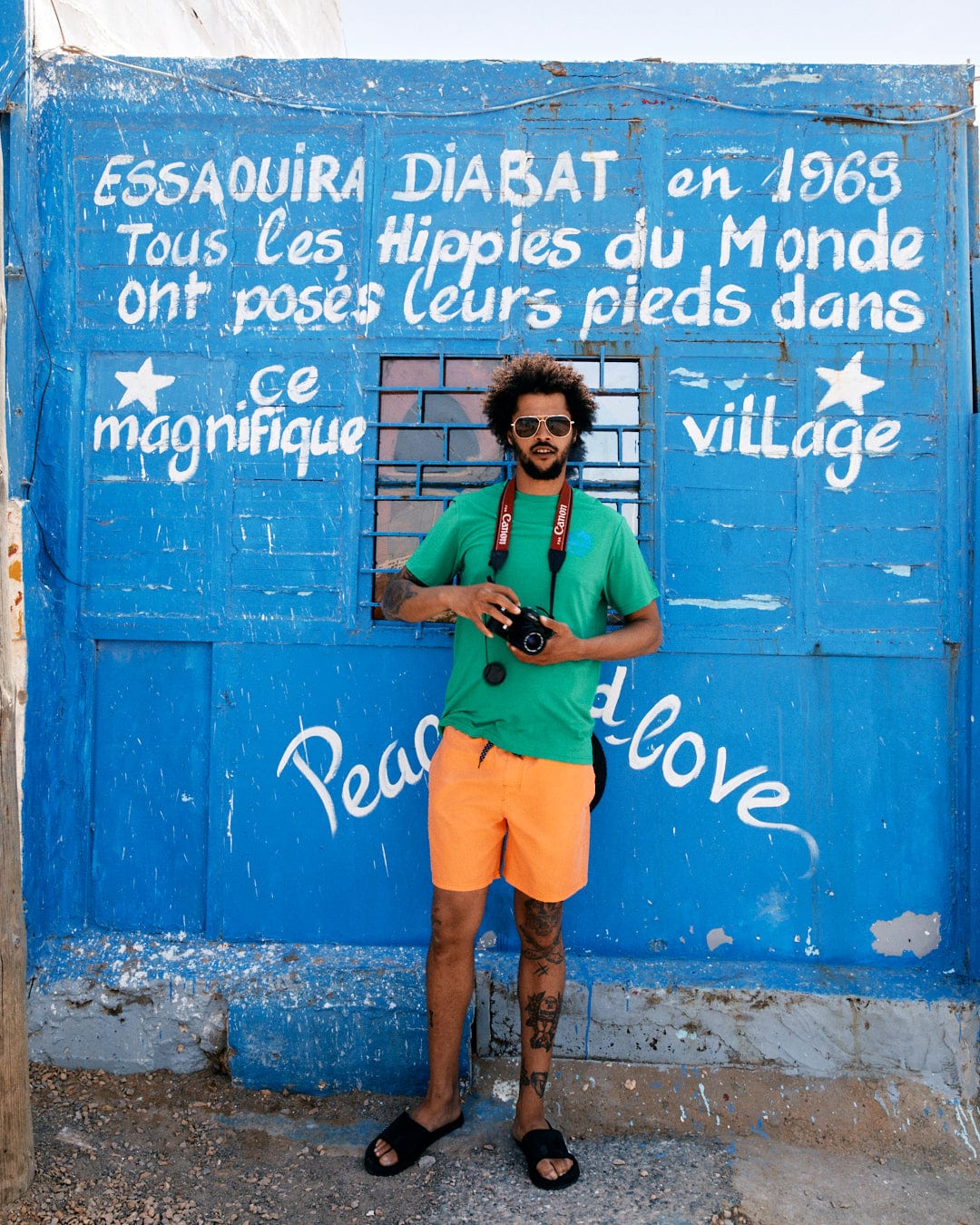 A man with a camera stands in front of a blue wall with French text about hippies in Essaouira Diabat, 1968. The wall, much like the Saltrock Geo Palms Solid - Mens Recycled Hybrid T-Shirt - Green, is adorned with the phrases 