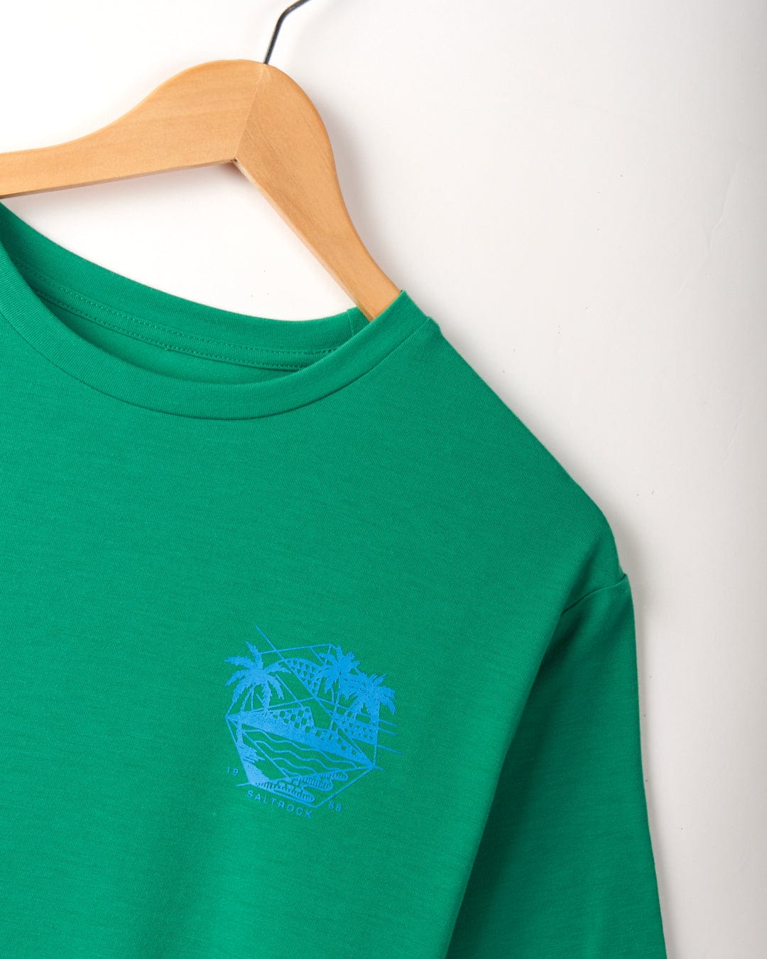 A Geo Palms Solid - Mens Recycled Hybrid T-Shirt - Green from Saltrock, featuring a blue Palm Beach graphic with palm trees and a hammock, made from recycled material, is hanging on a wooden hanger against a white background.