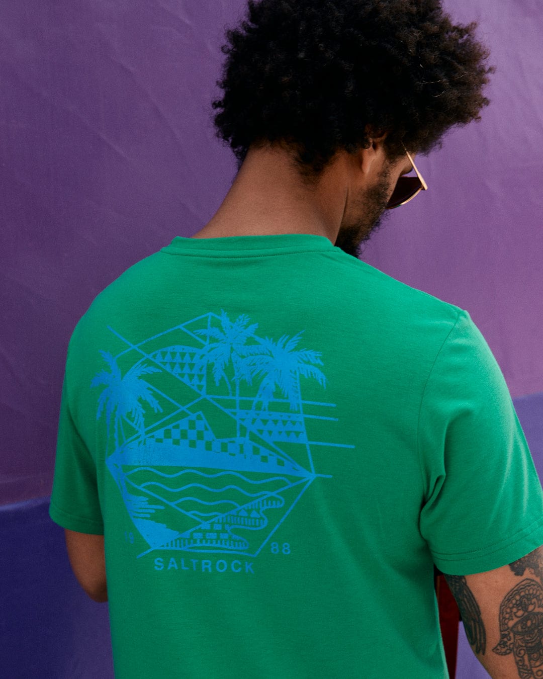 A person with curly hair, wearing sunglasses and a green Geo Palms Solid - Mens Recycled Hybrid T-Shirt - Green by Saltrock featuring Palm Beach graphics of palm trees and geometric shapes, stands in front of a purple wall, facing away from the camera.