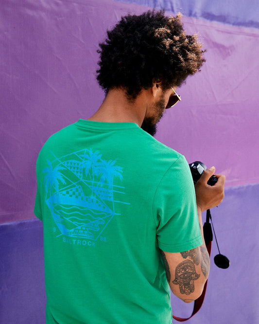 A person with curly hair and a tattooed arm, wearing a Geo Palms Solid - Mens Recycled T-Shirt - Green from Saltrock, is looking at a camera against a purple and pink background.