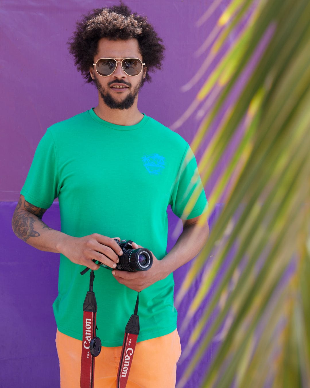 A man with curly hair wearing sunglasses, a Geo Palms Solid - Mens Recycled Hybrid T-Shirt - Green by Saltrock with a cotton soft hand feel, and orange shorts holds a Canon camera. A large palm leaf is in the foreground, evoking Palm beach graphics vibes. The background wall is purple.