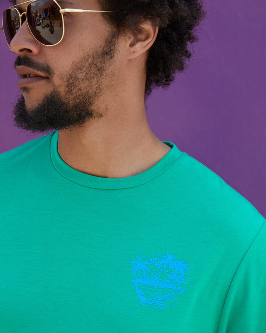 A man with curly hair, wearing sunglasses and a Saltrock Geo Palms Solid - Mens Recycled T-Shirt - Green, stands in front of a purple background.
