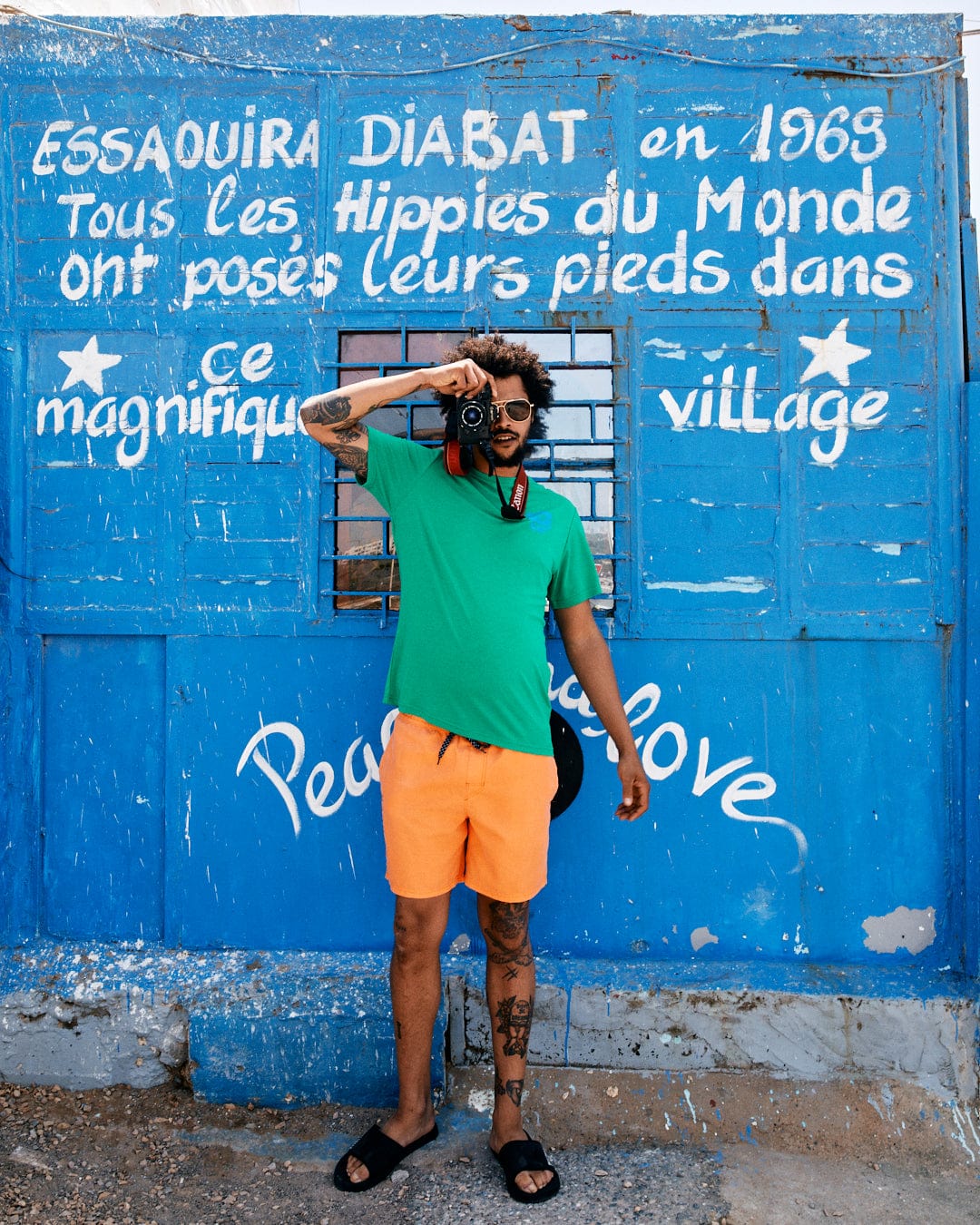 A person wearing the Geo Palms Solid - Mens Recycled T-Shirt - Green by Saltrock and orange shorts, made from recycled material, stands in front of a blue wall with white French text, holding a camera to their face. The wall features messages about hippies and peace and love. The outfit is stylish yet eco-friendly and machine washable.