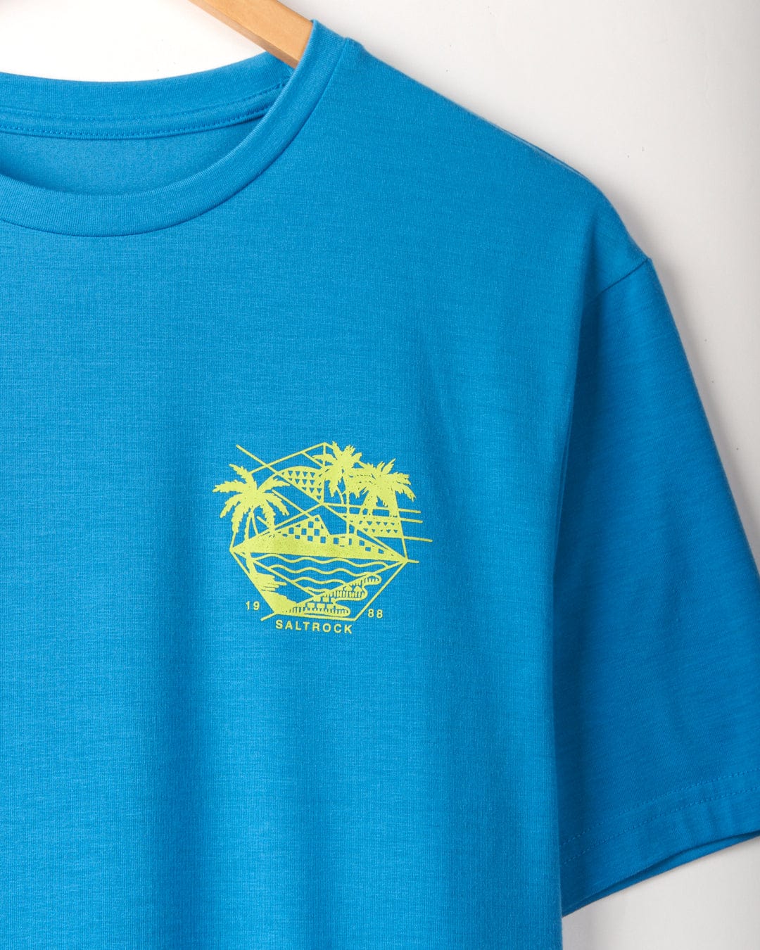 Geo Palms Solid - Mens Recycled T-Shirt - Blue hanging on a wooden hanger, featuring a yellow graphic design on the left chest depicting mountains, a river, and palm trees. Made from recycled material for an eco-friendly touch. The text "Saltrock 1988" is included in the design.