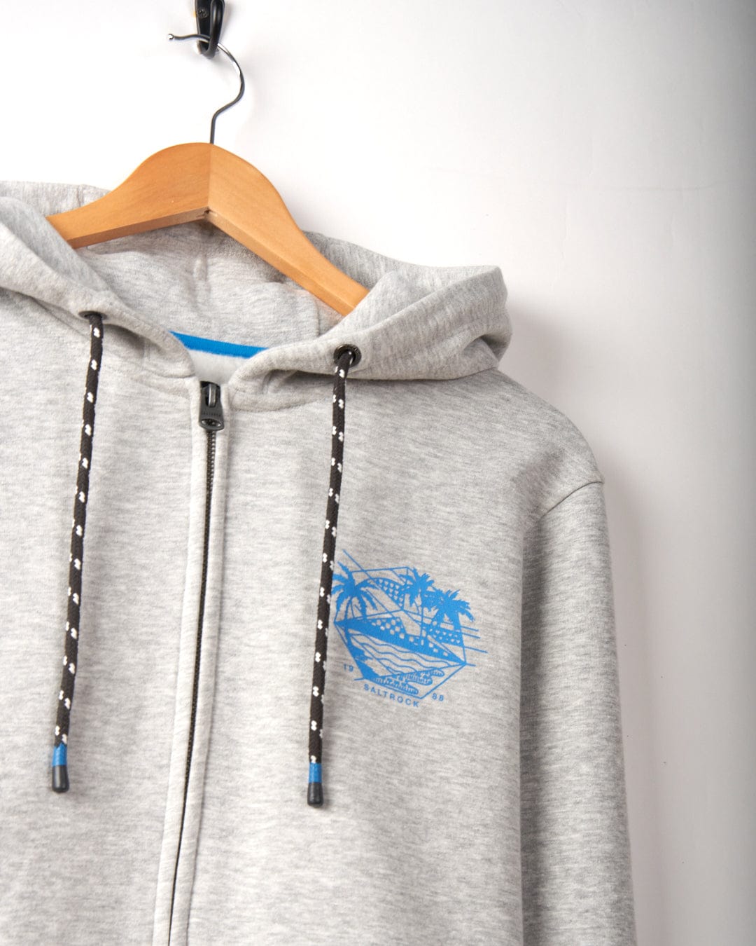 Gray hoodie with black and white drawstrings hanging on a wooden hanger. Blue graphic with palm trees and text on the left chest area features subtle Saltrock branding. Geo Palms - Mens Recycled Zip Hoodie - Grey on a white background.