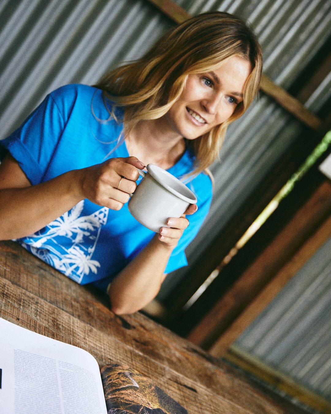 A woman wearing the Geo Beach Mono - Women's T-Shirt in blue and holding a white mug smiles at a wooden table with an open book, inside a corrugated metal structure featuring Saltrock branding.