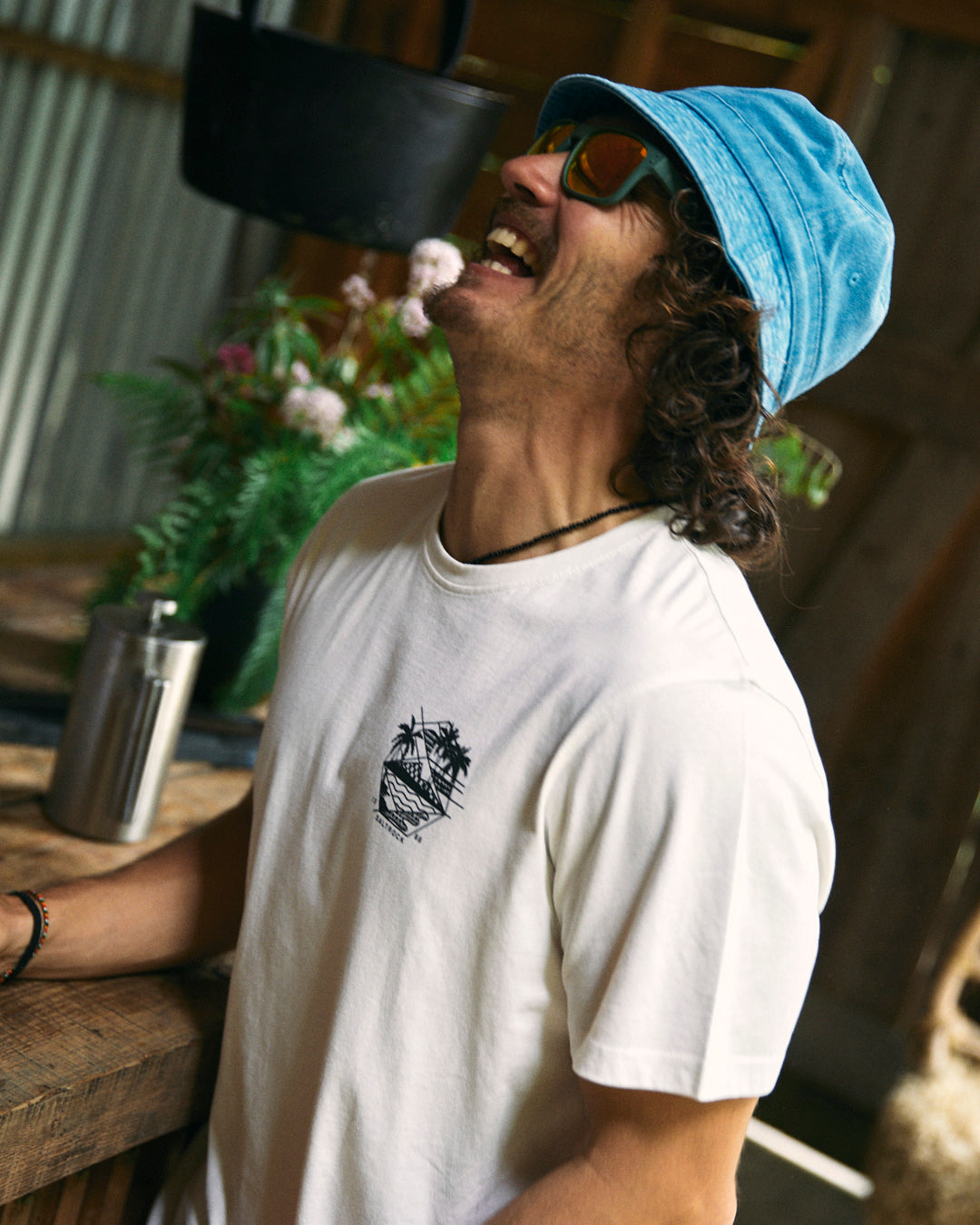 A man wearing sunglasses and a blue hat laughs while standing near a wooden counter with a metal bottle and flowers in the background. He is dressed in the white Saltrock Geo Palms Men's T-Shirt, made from 100% cotton.