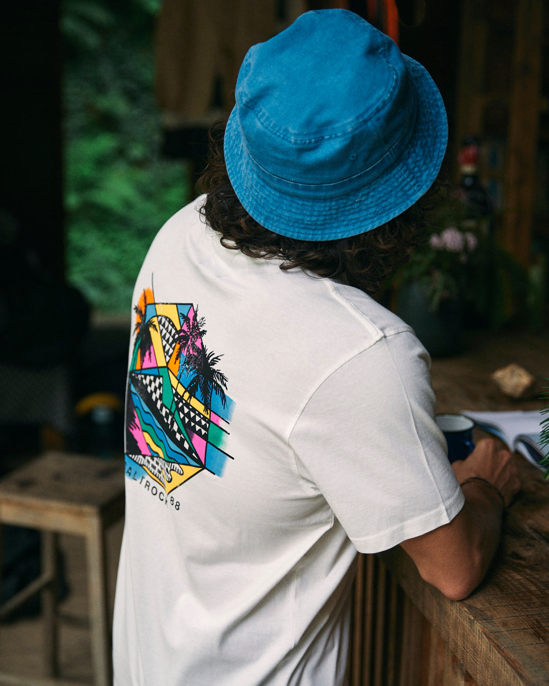 A person in a blue bucket hat with curly hair sits at a wooden table wearing the Geo Palms Men's T-Shirt from Saltrock, made of 100% cotton and featuring vibrant graphics of a sunset and palm trees on the back.