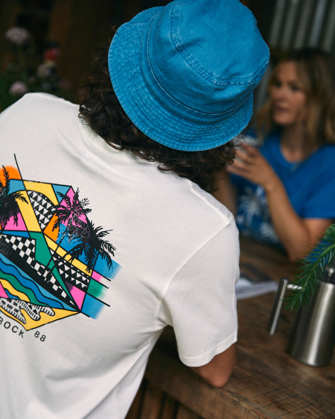 A person with long hair wearing a Saltrock Geo Palms men's white t-shirt featuring vibrant graphics sits at a wooden table, talking to another person in a blue shirt and blue hat.