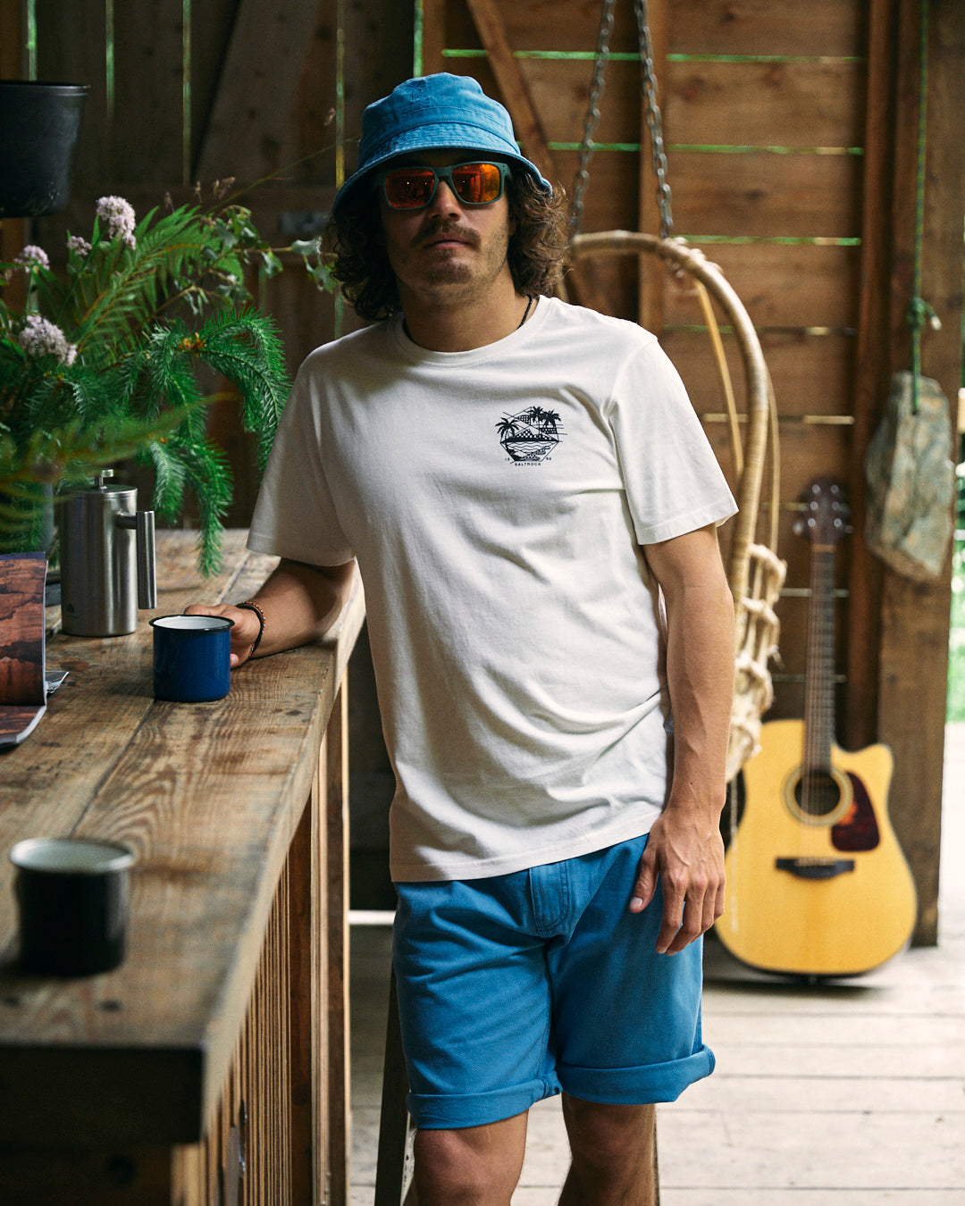 A person in a Geo Palms - Mens T-Shirt - White by Saltrock, blue shorts, and a blue hat stands indoors, leaning against a wooden counter with a blue mug in hand. A hanging chair, guitar, and plants are visible in the background.