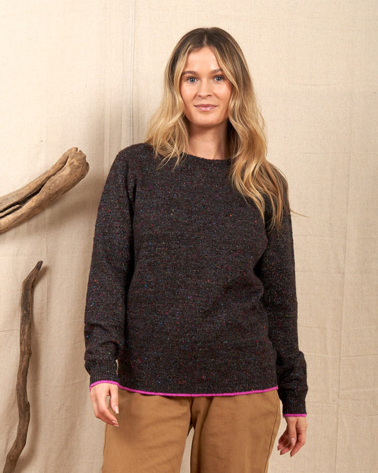 A person with long hair wears a Saltrock Gilly women's knitwear in dark grey, made from multi-color speckled yarn, paired with beige pants, standing indoors against a beige fabric backdrop with wooden branches.