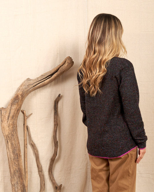 A person with long hair, wearing a Saltrock Gilly Women's Knitwear in dark grey and beige pants, stands with their back to the camera. Nearby, driftwood elements are artistically positioned against a plain backdrop.