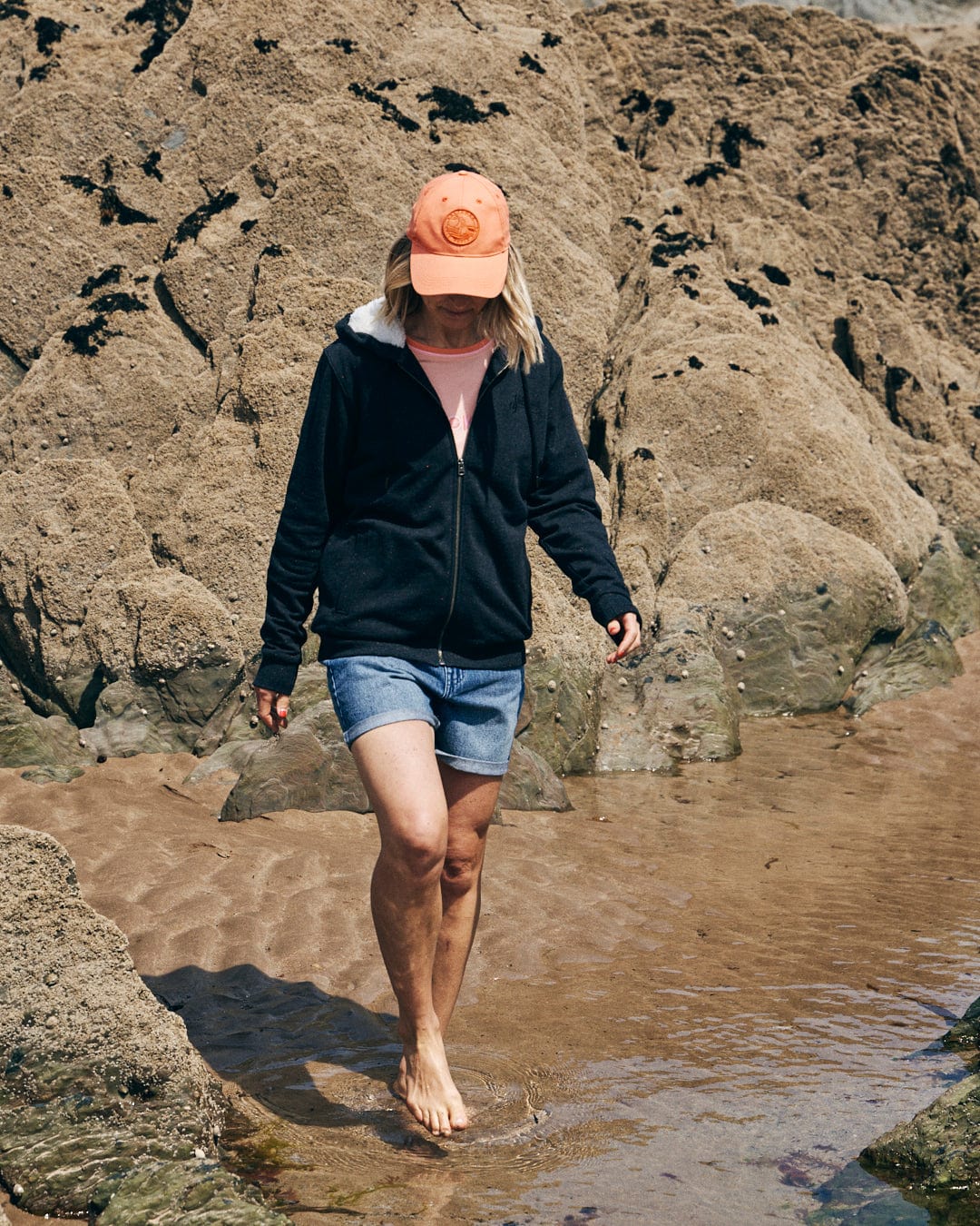 A person in a Grey Saltrock Ginny Women's Borg Lined Hoodie, denim shorts, and an orange cap walks barefoot on a rocky beach.