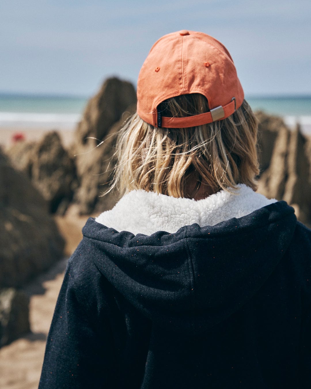 A person with shoulder-length blonde hair wearing an orange cap and a Ginny women's borg-lined hoodie in grey, featuring Saltrock branding, stands facing a rocky coastline.