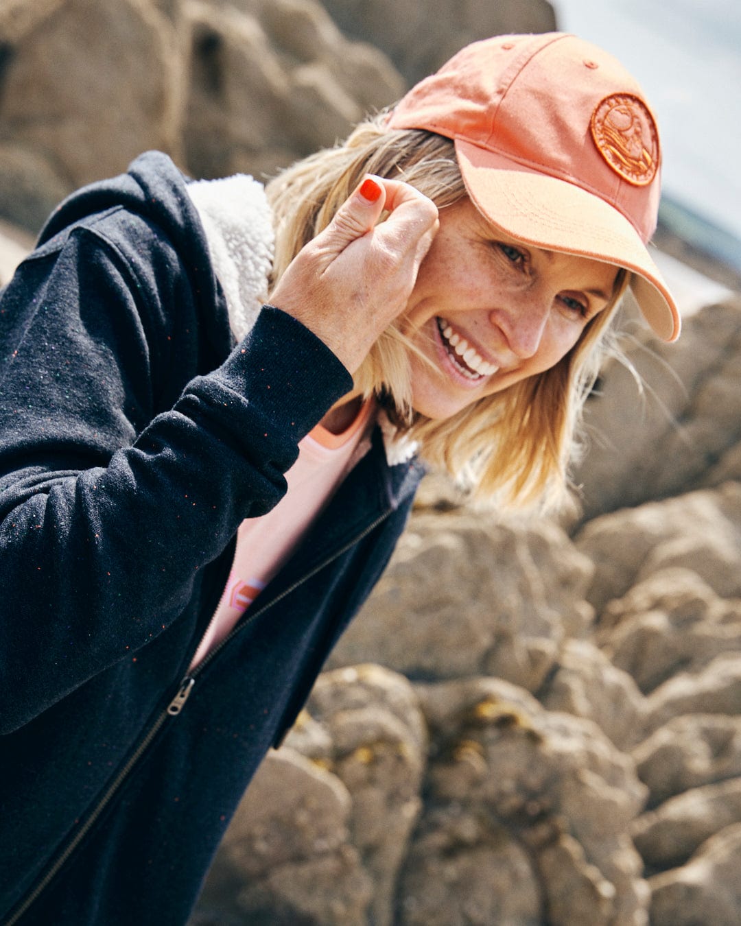 A person in a grey Saltrock Ginny Borg lined hoodie with an orange cap smiles while standing outdoors near rocky terrain.