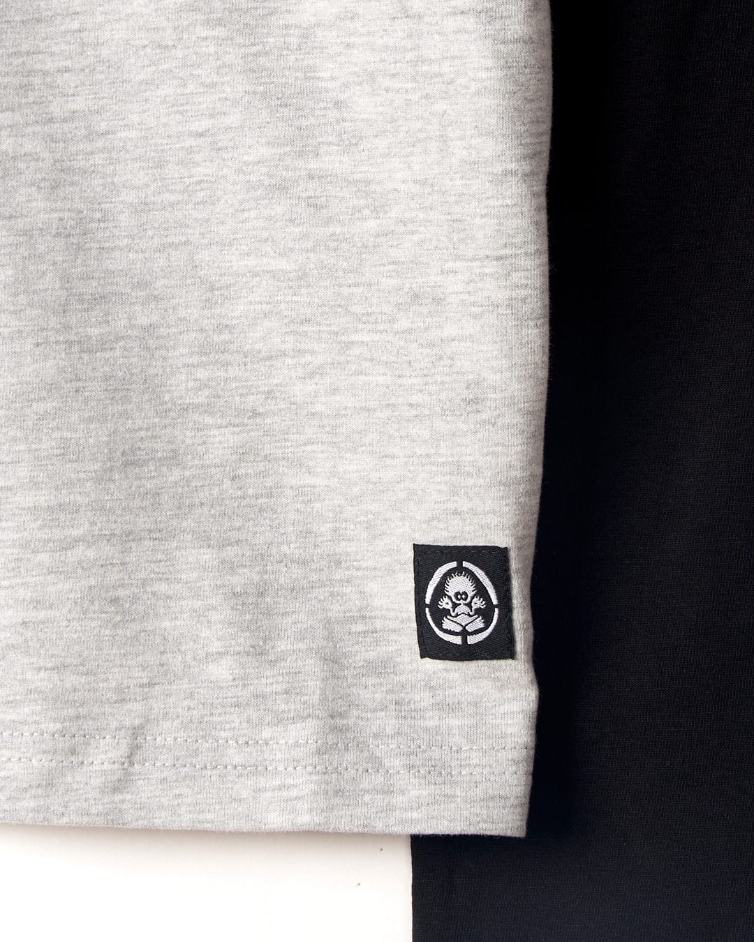 Close-up of a gray fabric with a small black and white Saltrock logo featuring a skull and crossbones near the bottom edge. The fabric is positioned next to a black fabric with raglan contrast sleeves. The garment, known as the Hardskate Warp - Kids Longsleeve Raglan T-Shirt in Grey/Black by Saltrock, is machine washable for easy care.