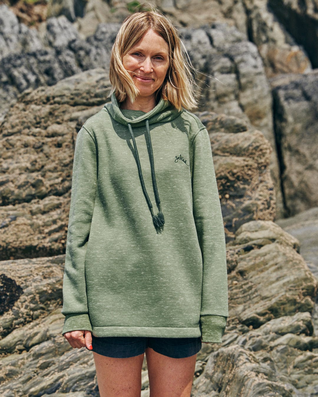 A person stands outdoors in front of large rocks, wearing the Saltrock Harper - Women's Longline Pop Sweat in green and shorts, their hands by their sides.