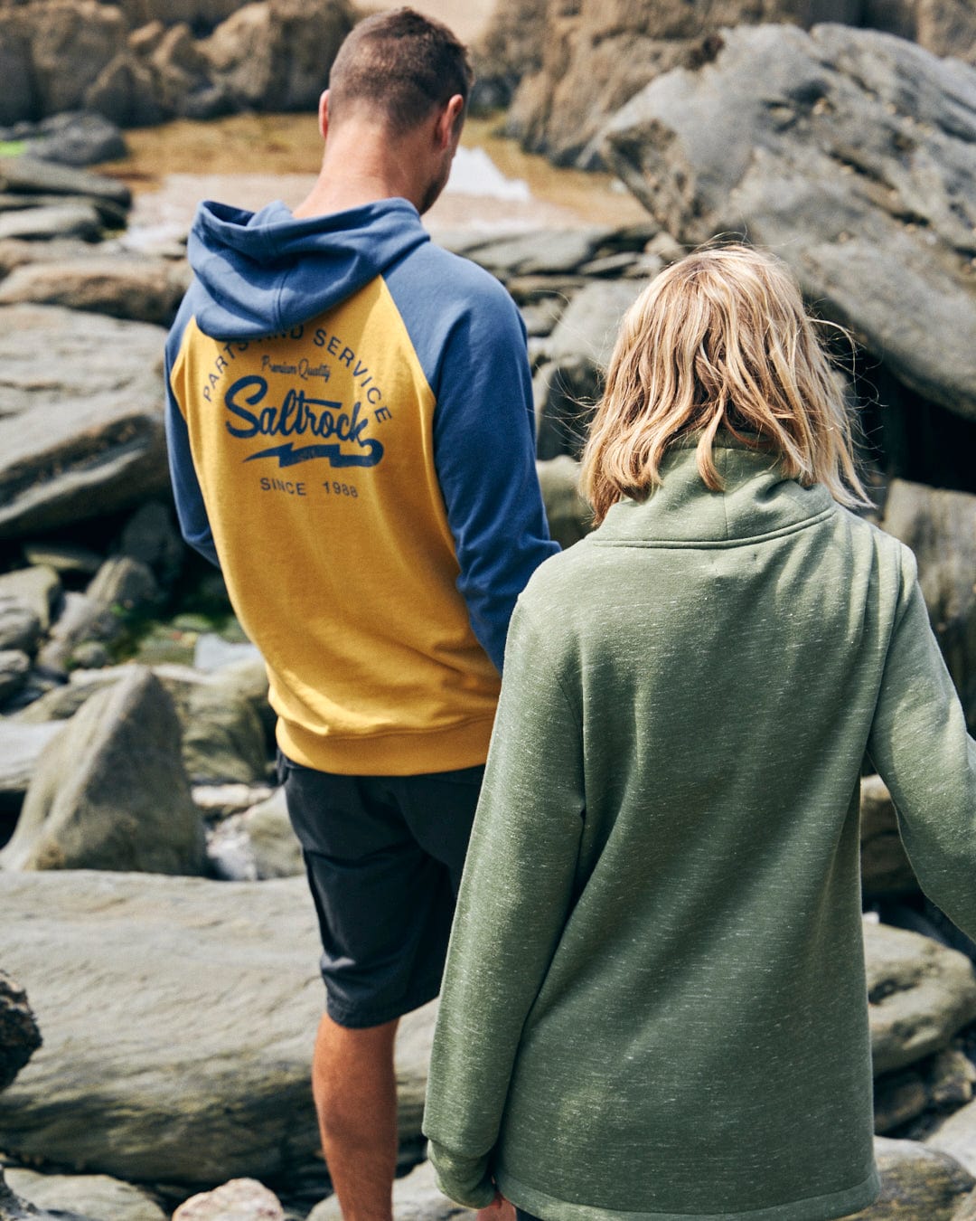 Two individuals in casual attire walk together on a rocky beach. The person on the left wears a yellow and blue hoodie with embroidered branding, while the person on the right sports the Saltrock Harper Women's Longline Pop Sweat in green.