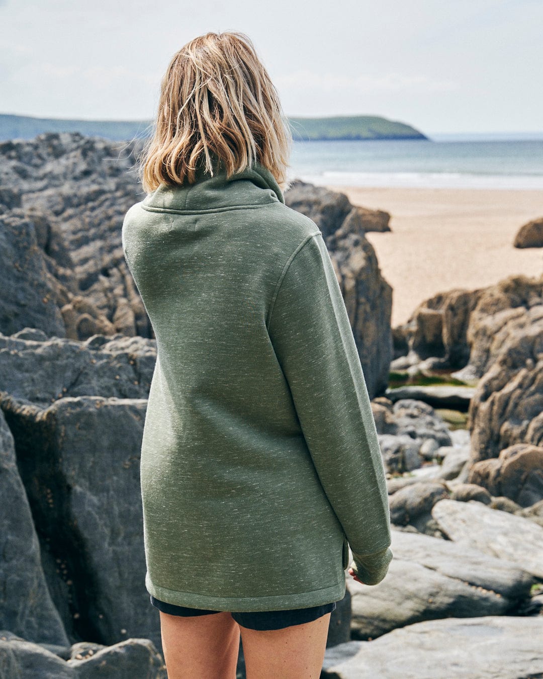 A person with shoulder-length blond hair, wearing the Saltrock Harper - Women's Longline Pop Sweat in green and shorts, stands on rocky terrain looking towards a distant sandy beach and ocean.