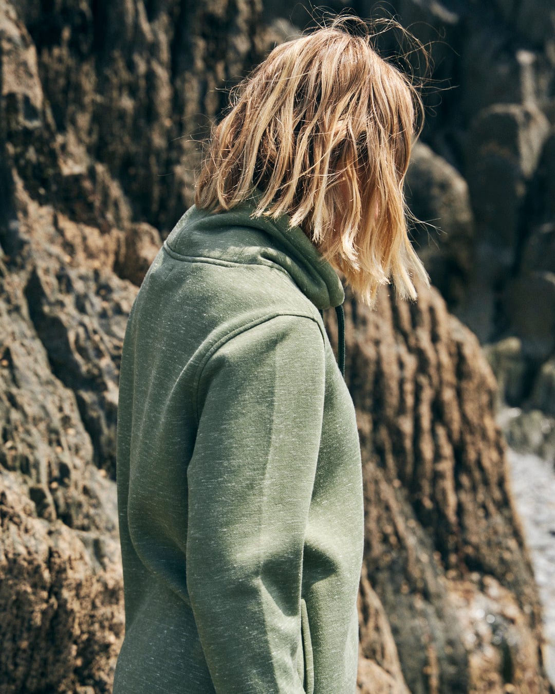 Person with shoulder-length blond hair is wearing the Saltrock Harper Women's Longline Pop Sweat in green and standing near large rocks.