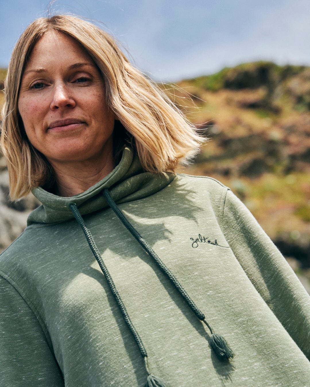 A person with shoulder-length hair is wearing the Saltrock Harper Women's Longline Pop Sweat in green, featuring embroidered branding, and is standing outdoors with a blurred natural background.