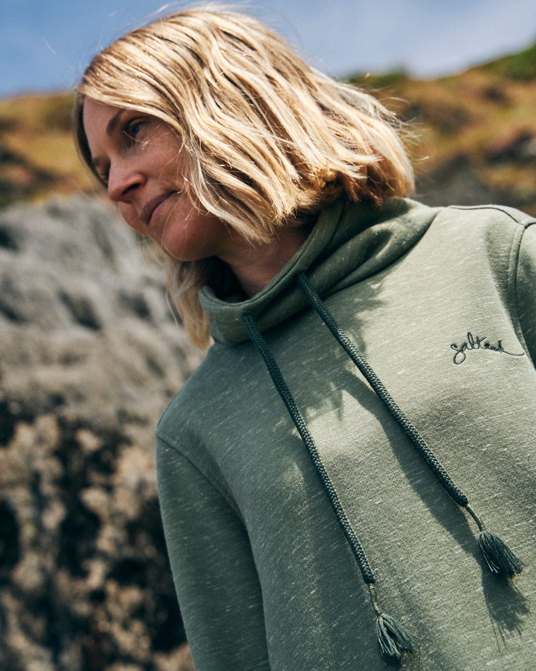 A person with shoulder-length blonde hair is wearing the Harper - Womens Longline Pop Sweat by Saltrock, featuring a green color, drawstrings, and embroidered branding, standing outdoors in front of rocky terrain.